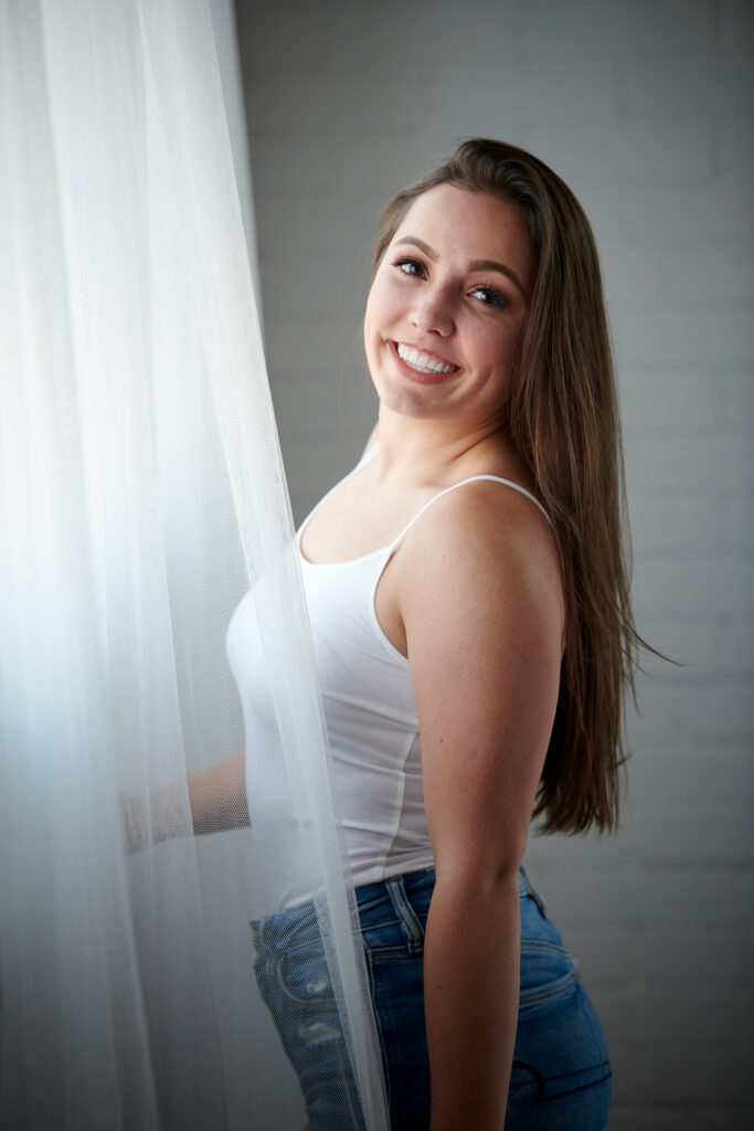 Sexy young brunette woman in white tank top and blue jeans stands near window